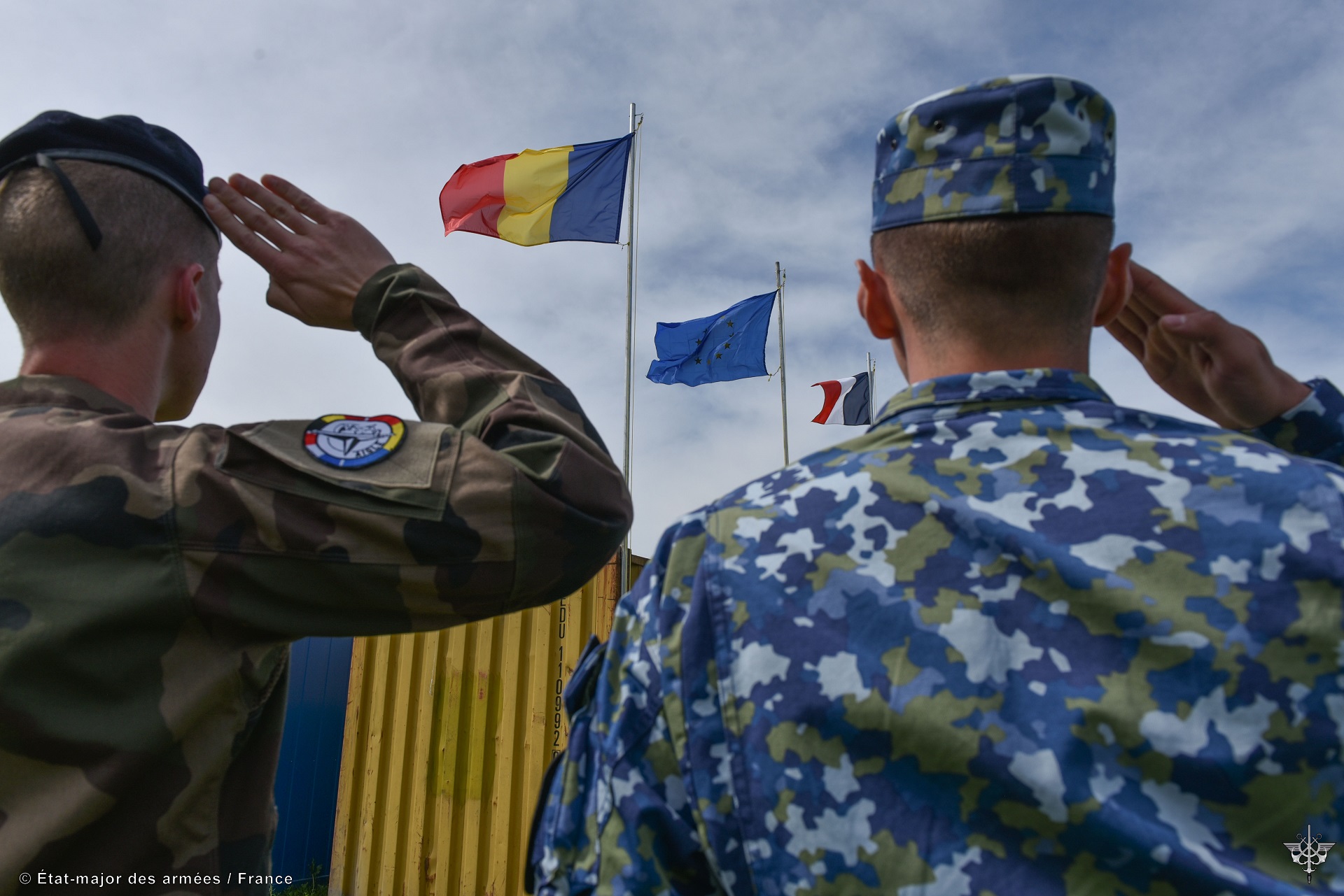 Un officier français et un officier roumain saluent les drapeaux roumain,européen et français.